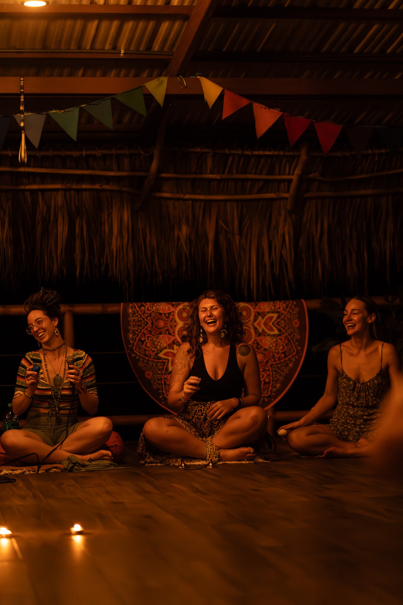 Three women sitting on the floor, laughing and smiling, with a banner of flags and candles, representing the joyful and communal atmosphere of the "Sol y Luna" retreat.